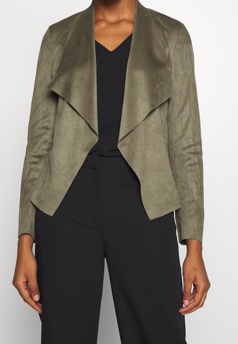 Blazer in ecopelle scamosciata verde oliva con apertura frontale e colletto sceso. Caratterizzato da maniche lunghe e una vestibilità rilassata. Indossato sopra un outfit nero.