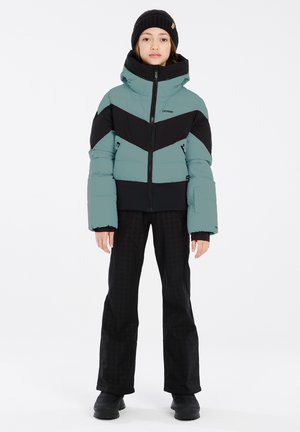 Jeune personne portant un bonnet noir, une doudoune bleu sarcelle et noire, un pantalon noir et des bottes noires, debout sur fond blanc.