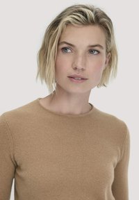 Blonde vrouw met kort haar, gekleed in een lichtbruine lange mouw sweater, kijkt iets naar rechts tegen een effen achtergrond.