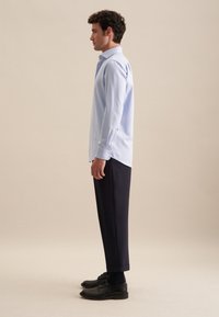 Camicia azzurro chiaro con un motivo sottile, abbinata a pantaloni blu scuro e scarpe nere, vista di lato.