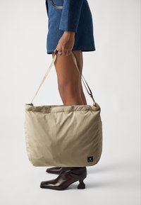Beige nylon toteveske med en jevn tekstur, glidelås, og lange, mønstrede stropper, holdt av en person iført en blå drakt og brune sko.