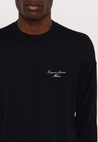 Homme portant un pull noir à col rond avec le logo blanc en cursive "Emporio Armani Milano" sur la poitrine gauche.