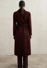 Burgundy uldfrakke med bælte i taljen, lange ærmer og sideslidser. Skræddersyet design med knapdetaljer på ærmeenderne.