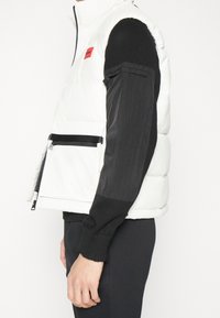 Weißer Puffer-Weste mit hohem Kragen, schwarzen Ärmeln, rotem Logo-Patch und einer Reißverschlusstasche auf der Vorderseite, mit strukturierten Stoffdetails.