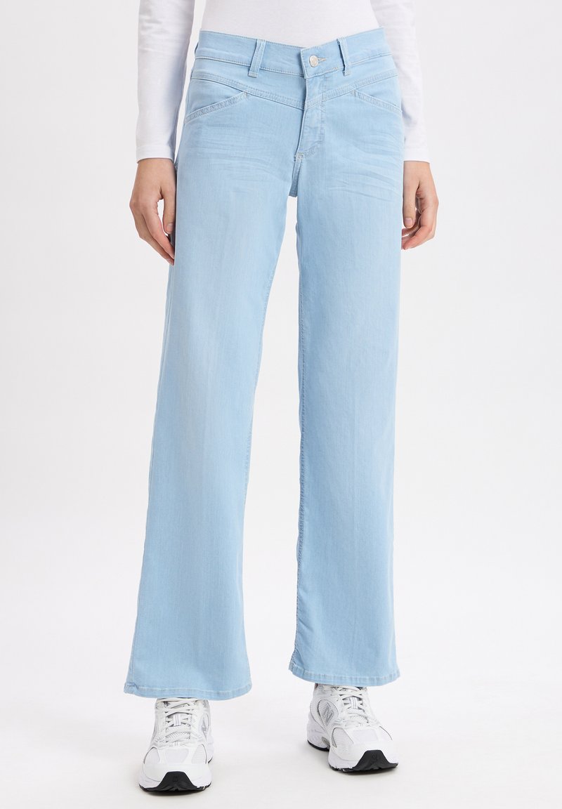 Hellblaue weit geschnittene Jeans aus Denim, mit Knopfverschluss, Fronttaschen und einer glatten Textur. Kombiniert mit weißen Sneakers.