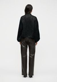 Γυναίκα όρθια με την πλάτη στραμμένη, φοράει μαύρο, υφασμάτινο πουκάμισο με μακριά μανίκια και σκούρα καφέ δερμάτινο παντελόνι ίσιας γραμμής, συνδυασμένο με μαύρες γόβες.