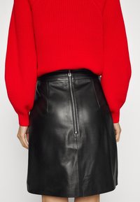 Vue de dos d'une personne portant un pull en tricot rouge et une jupe en cuir noir avec une fermeture éclair argentée visible à l'arrière.