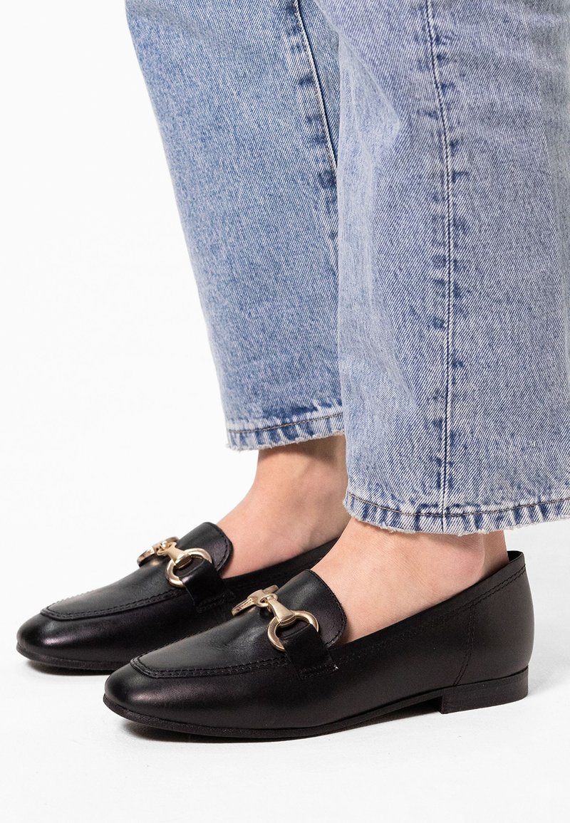 Black leather loafers with a rounded toe design, featuring a prominent gold-tone hardware accent and a low flat heel. Worn with light blue jeans.