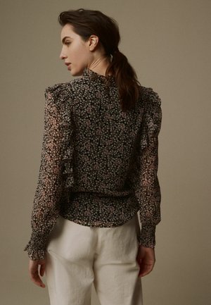 Schwarze Blumenbluse aus durchscheinendem Stoff, mit volantartigen Schultern und langen Ärmeln. Das Muster zeigt kleine rosa und weiße Blumen. Weiße Hosen sind sichtbar.