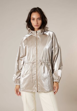 Vrouw draagt een metallic beige capuchonjas met een voorrits en een trekkoord in de taille, gecombineerd met off-white broek, staand tegen een effen achtergrond.