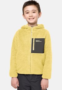Keltainen fleece-takki hupulla, jossa on musta vetoketjullinen rintatasku ja kevyet vetoketjuakcentit. Pehmeä rakenne, rento muotoilu.