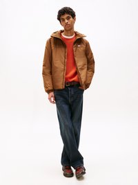 Giacca cappuccio marrone con zip frontale, maglione arancione, jeans scuri e sneakers con motivo. Il modello sta dritto su uno sfondo neutro.