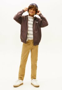 Sudadera morada con bolsillos frontales sobre una camiseta de rayas, combinada con pantalones beige y zapatillas blancas, con un gorro azul en la cabeza.