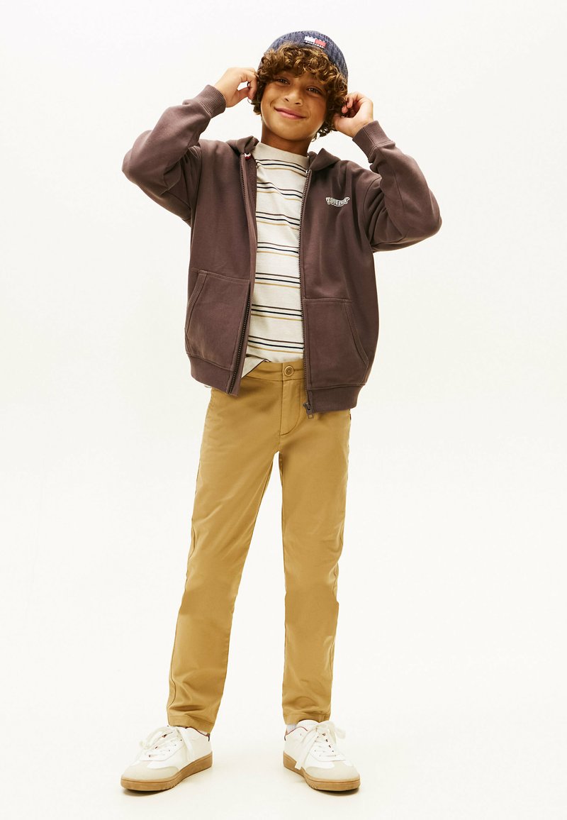 Sudadera morada con bolsillos frontales sobre una camiseta de rayas, combinada con pantalones beige y zapatillas blancas, con un gorro azul en la cabeza.