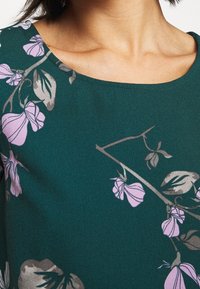 Tessuto stampato floreale verde con fiori rosa e foglie grigie, con una trama liscia e un collo arrotondato.