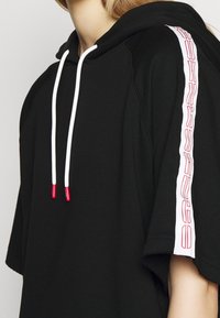 Sort hættetrøje med korte ærmer, med hvide snore med røde ender og en hvid lodret logoaccents på ærmet.