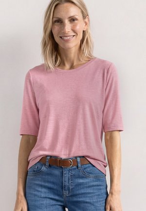 Uśmiechnięta kobieta o blond włosach nosi różową koszulkę z krótkim rękawem wpuszczoną w niebieskie dżinsy z brązowym paskiem, stoi na jednolitym tle.