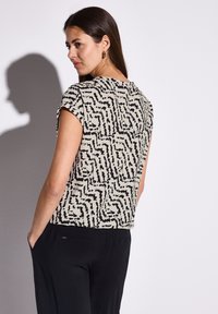 Maglietta a maniche corte in un tessuto beige con un motivo astratto nero. Presenta una vestibilità rilassata e un collo rotondo; abbinata a pantaloni neri.