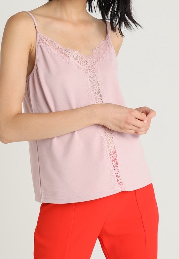 Femme portant un top sans manches rose clair bordé de dentelle, associé à un pantalon rouge vif, mains légèrement croisées à la taille.
