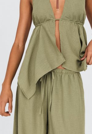 Woman wearing loose, light olive green sleeveless top with deep V-neck and matching drawstring pants against plain background.