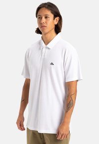 Weißes Polohemd mit Kragen und kurzen Ärmeln, versehen mit einem kleinen schwarzen Logo auf der Brust, aus weichem, strukturiertem Stoff.
