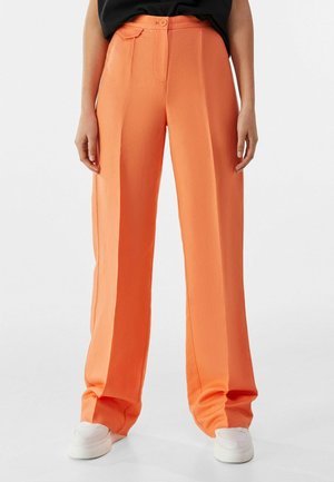 Forfra af en person iført orange bukser med høj talje og vide ben, kombineret med hvide sneakers og en sort top, stående mod en ensfarvet baggrund.