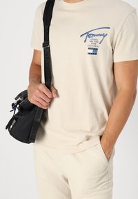 Beige bavlněné tričko s modrým logem na hrudi, v kombinaci se světle krémovými kalhotami. Malá černá crossbody taška s nastavitelným popruhem.