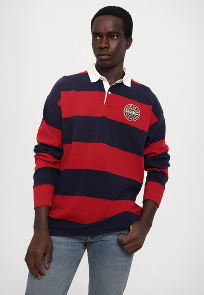 Tricou de rugby cu dungi roșii și bleumarin, cu guler alb, având o aplicație brodată circulară pe piept și mansete ribbed.