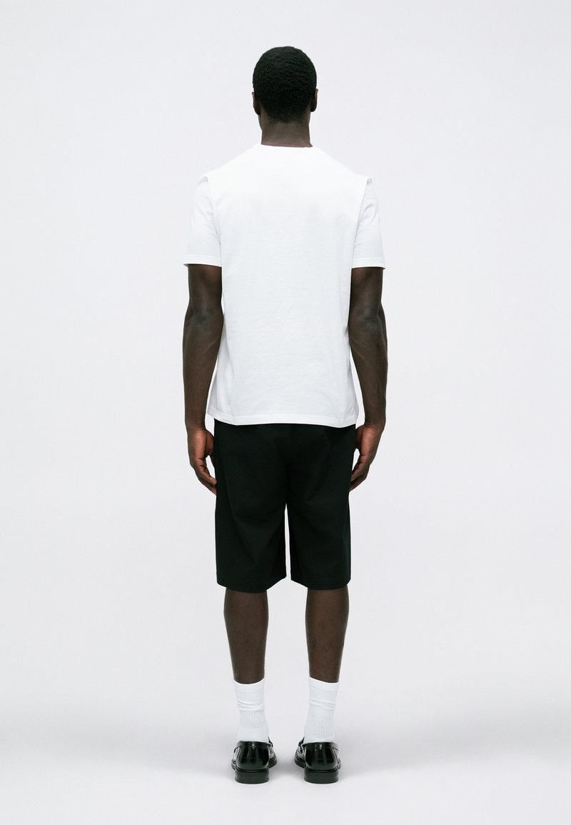 Witte t-shirt met korte mouwen, zwarte shorts tot op de knie en zwarte schoenen. Model staat met de rug naar de camera tegen een eenvoudige lichtkleurige achtergrond.