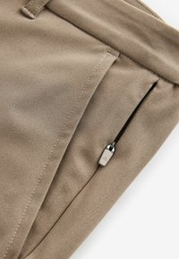 Beige Chinos mit einer glatten Stofftextur, einem gefalteten Vorderteil und einer Reißverschlusstasche mit einem dreieckigen Metallreißverschlussanhänger.