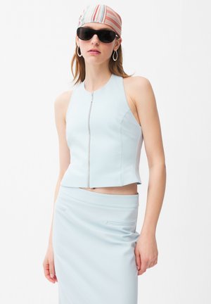 Kobieta ubrana w jasnoniebieski top bez rękawów na zamek błyskawiczny oraz dopasowaną spódnicę, w paskowaną chustę na głowę, duże kolczyki koła i czarne okulary przeciwsłoneczne.