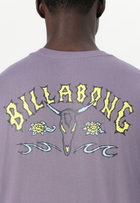 Lilla bomulls-T-skjorte med fet gul 'BILLABONG'-bokstaver, en stilisert hodeskalle, blomster og bølgemønster på baksiden.