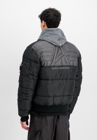 Chaqueta acolchada negra con una sección superior gris y capucha gris, con puños y cinturilla acanalados, y un bolsillo lateral con cremallera.