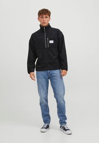 Pulover negru din fleece cu guler înalt, fermoar pe jumătate în față și buzunar pe piept; asortat cu blugi albastru deschis și teneși negri.