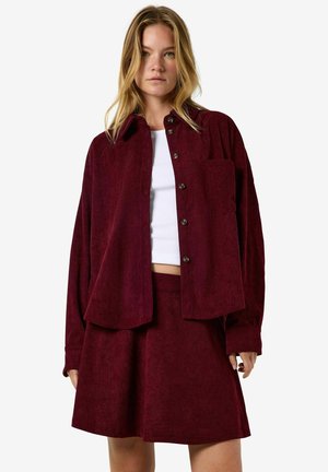 Femme portant une veste boutonnée en velours côtelé rouge foncé et une jupe assortie avec un crop top blanc en dessous, sur fond blanc.