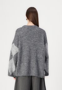 Nainen, jolla on pitkät ruskeat hiukset, pukeutuneena tilavaan harmaaseen neulepuseroon, jossa on vaaleita ja tummia timanttimaisia kuvioita hihoissa, yhdistettynä harmaisiin housuihin, takanäkymä.