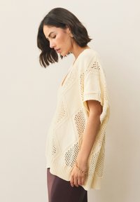 Pleteni kremno-bel pulover z vzorcem diamantov, kratkimi rokavi in rebrastim robom, nošen čez temne hlače. Poudarjen z zlatim zapestnim trakom.