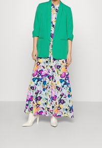 Světlý zelený blazer přes maxi šaty s květinovým potiskem, který obsahuje růžové, fialové a modré akcenty. Model nosí bílé špičaté podpatky.