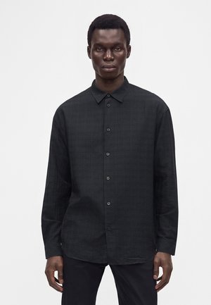 Homme portant une chemise noire à manches longues avec un motif à carreaux subtil, debout devant un fond blanc uni.