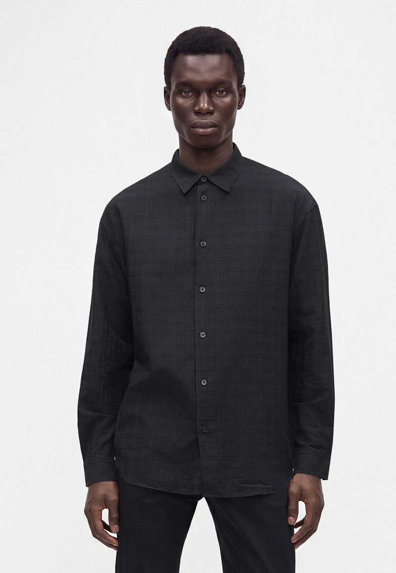 Homme portant une chemise noire à manches longues avec un motif à carreaux subtil, debout devant un fond blanc uni.