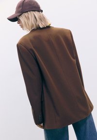 Brun blazer med en glat tekstur, enkelt bagskær, og fire-knap mansjettdetalje, parret med denim bukser og en brun kasket.