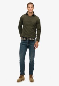 Oliwkowy sweter z wytłoczonym tekstem "Superdry Athletics", nałożony na koszulę w kratę, w połączeniu z dżinsami i brązowymi butami.