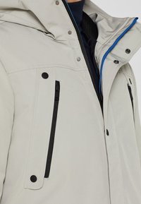 Giacca grigia chiara con chiusura a zip, accessori neri e accenti blu. Presenta due tasche sul petto e un rivestimento interno testurizzato.