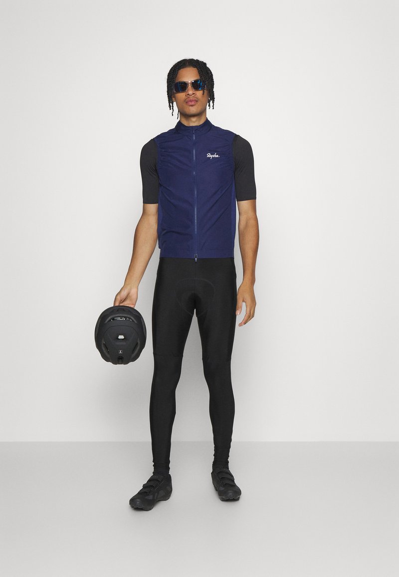 Marineblauw mouwloos fietsvest, zwarte strakke leggings, zwarte fietsschoenen, houdt een zwarte helm vast en draagt een zonnebril. Glad stoffen textuur.