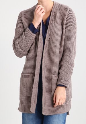 Femme portant un long cardigan taupe en maille épaisse avec des poches avant, par-dessus un haut bleu marine et un jean bleu, touchant son menton et une poche.