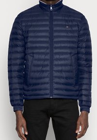 Granatowa kurtka puchowa z zamkiem błyskawicznym, stojącym kołnierzem i wieloma poziomymi pikowanymi sekcjami. Posiada boczne kieszenie i elastyczne mankiety.