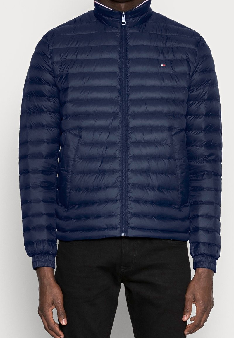 Granatowa kurtka puchowa z zamkiem błyskawicznym, stojącym kołnierzem i wieloma poziomymi pikowanymi sekcjami. Posiada boczne kieszenie i elastyczne mankiety.