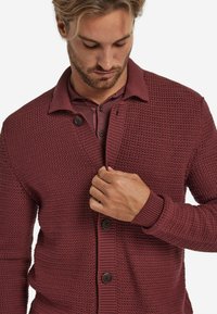 Mörkröd stickad cardigan med ett strukturerat mönster, försedd med krage, knappstängning och ribbade manschetter. Bärs över en matchande skjorta.