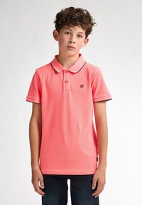 Korálový polo top s límcem, dvěma knoflíky a kontrastním tmavým lemem. Jemná, hladká textura, spárována s tmavými džínami.