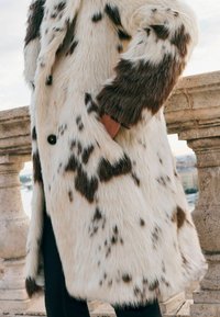 Umělá kožešina v krémové a hnědé barvě s tečkovaným vzorem. Obsahuje límec, boční kapsy a zapínání na knoflíky. Měkká textura.